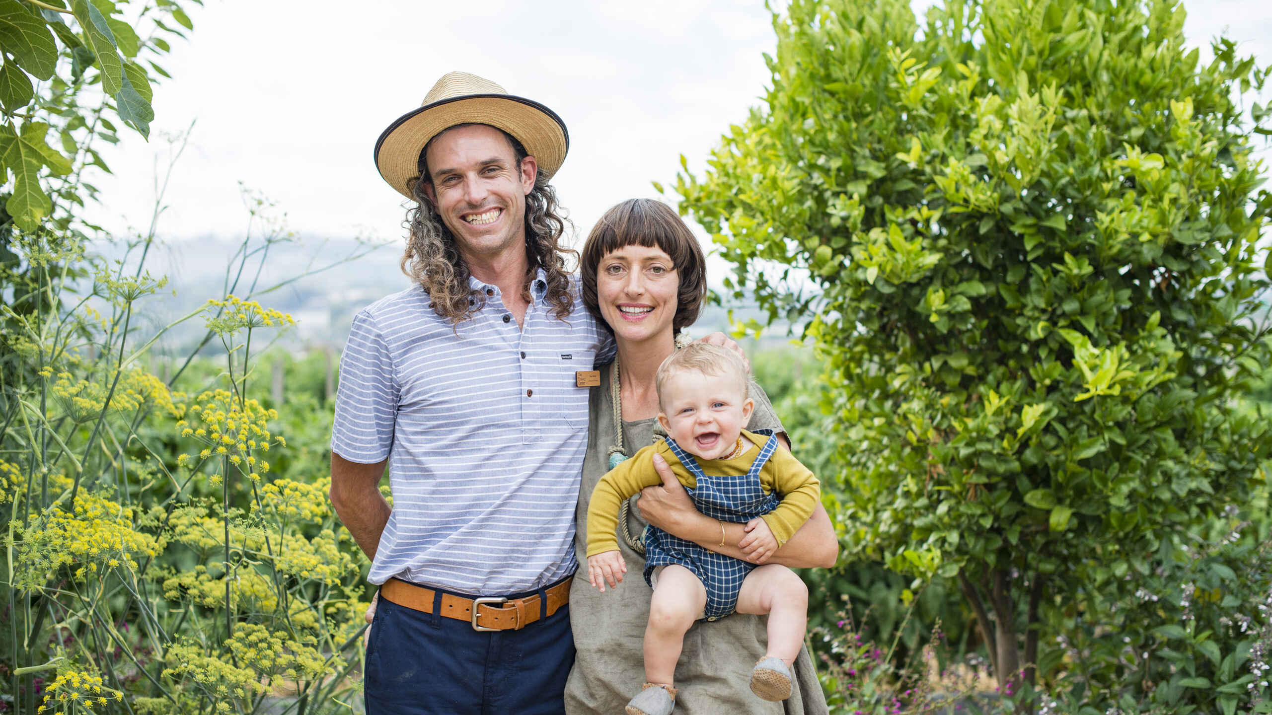 A farmer and woman with their young child stand together in a lush vegetable garden