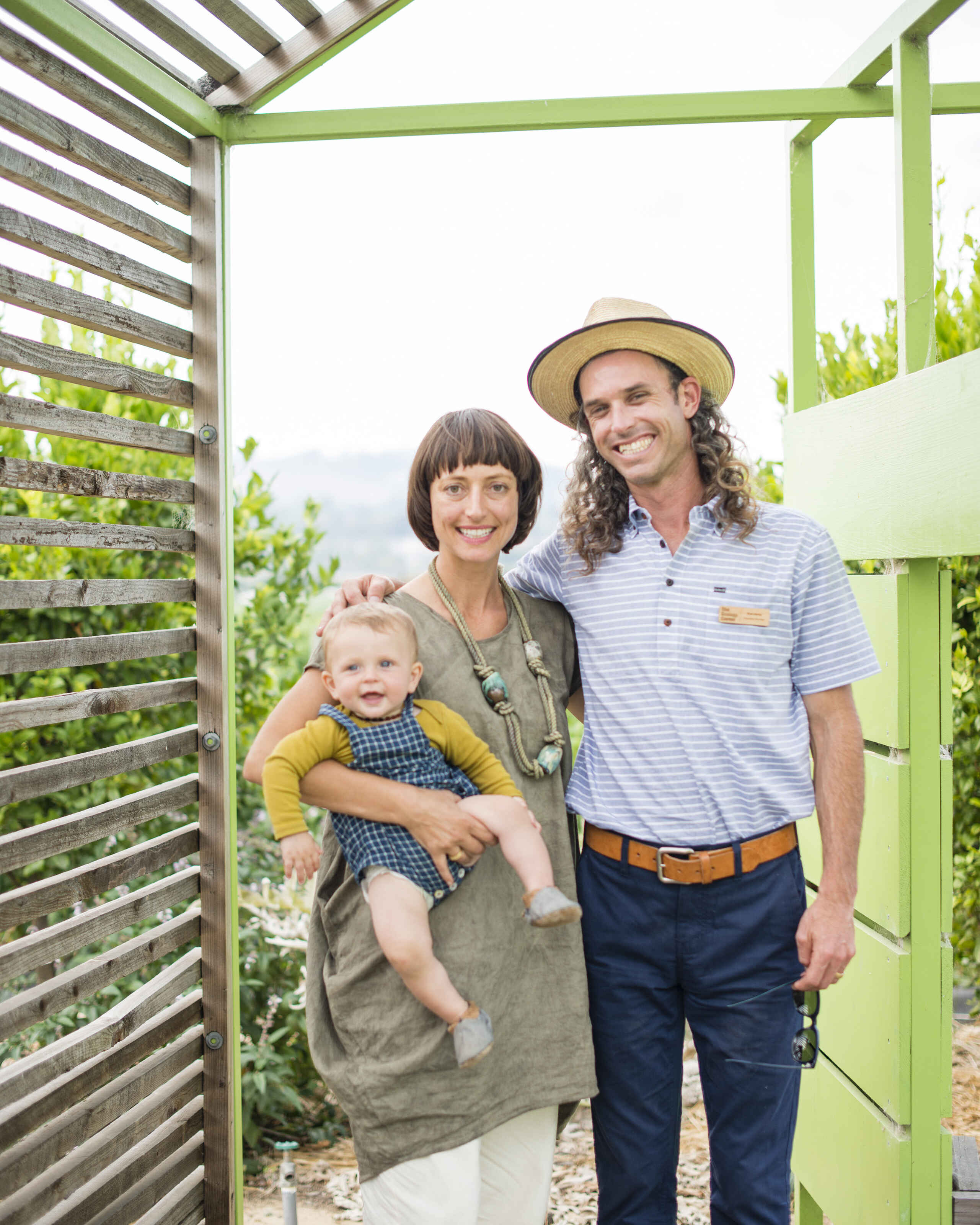 A family of three stands together in a green garden structure, smiling at the camera
