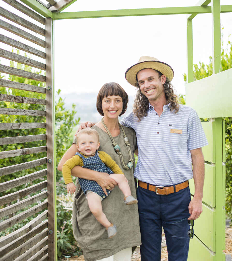A family of three stands together in a green garden structure, smiling at the camera