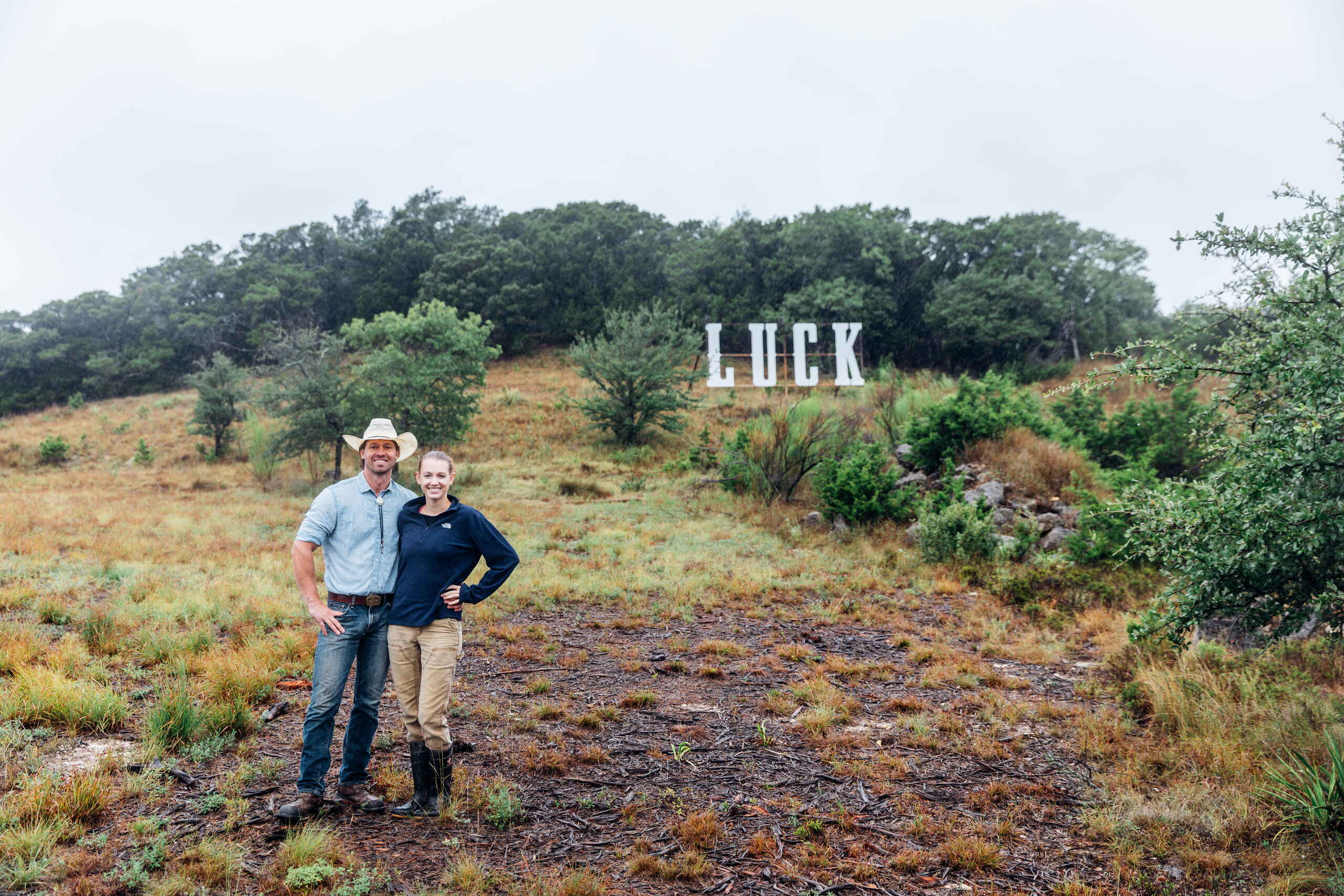 Tina y Orion Weldon de pie en sus tierras de rancho con el letrero Huck visible en la ladera detr&aacute;s de ellos