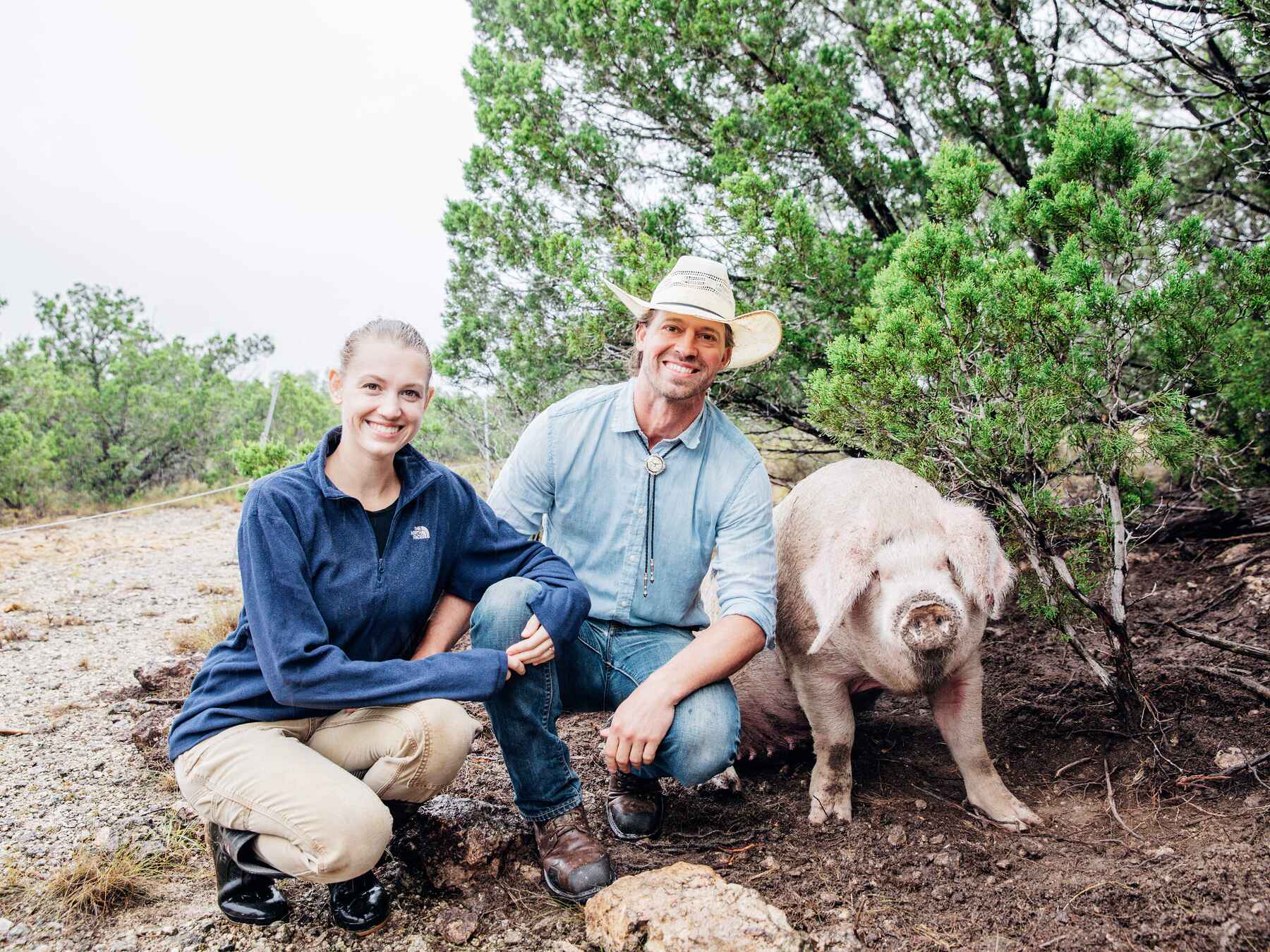 Tina and Orion Weldon crouch beside their pig on a ranch trail surrounded by trees