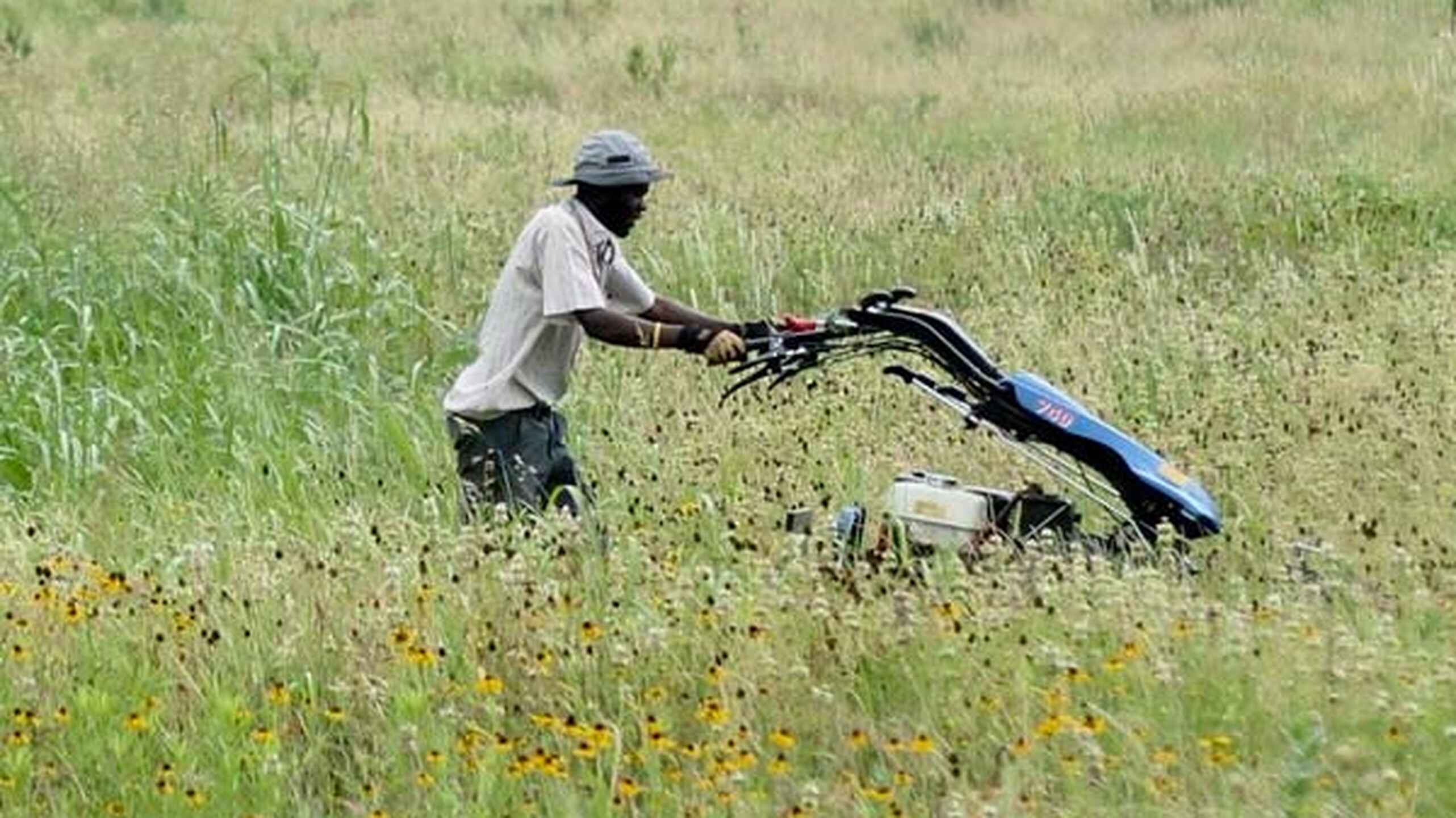 Wandaka Heritier Musungira pushes a blue motorcycle through a wildflower-dotted pasture toward tree-lined fields