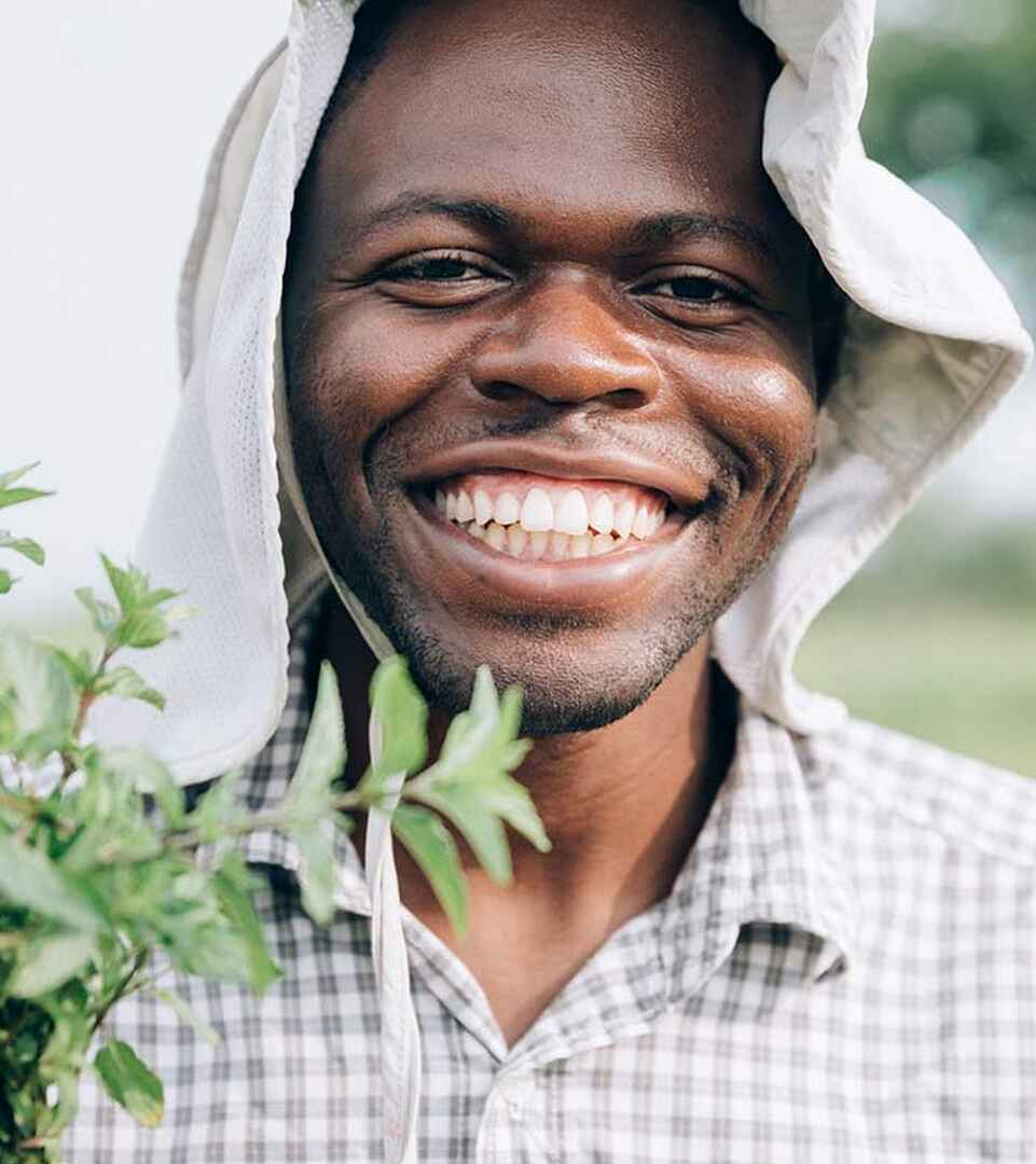 Wandaka Heritier Musungira smiling while holding a seedling, wearing a white head wrap and checkered shirt