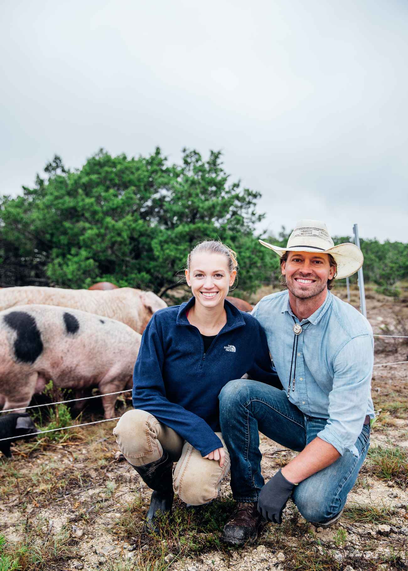A farmer and young woman crouch together in front of grazing pigs on their ranch